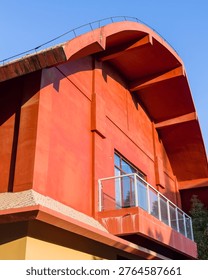 Abstract low angle perspective of a vibrant red building exterior featuring a dramatic curved roof and glass balcony under a blue sky. The scene represents creative architectural forms, modern educational campuses, and bold urban design.