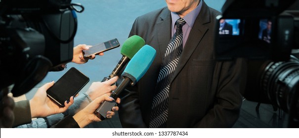 Abstract Man In A Suit And Tie Speaks To Reporters And Video Cameras. Female Hands Hold Microphones, Voice Recorders And Mobile Phones To Record Sound. Interview, Report, Media.