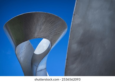 Abstract metal sculpture with twisting, open form against a bright blue sky. Smooth, reflective surface and dynamic curves ideal for modern art, architecture, and urban themes. - Powered by Shutterstock