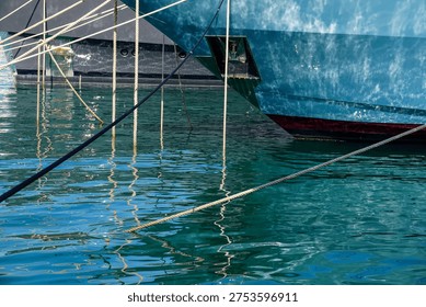 Abstract nautical composition featuring the bow of a luxury superyacht moored in a harbor. The image showcases deep turquoise sea water with intricate ripples and reflections of the sunlight hitting the yacht's hull. Several thick mooring ropes cut through the frame, creating strong diagonal lines that add a sense of perspective and industrial elegance. This high-quality commercial photograph, taken in a Mediterranean marina like Porto Montenegro, perfectly illustrates themes of maritime safety, luxury travel, yachting lifestyle, and port infrastructure. The vibrant blue and teal tones make it ideal for marine industry brochures, web backgrounds, and transport-themed marketing.