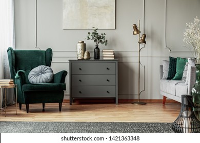 Abstract painting on grey wall o contemporary living room interior with emerald green armchair with round pillow, commode and couch