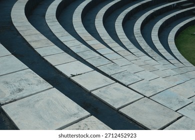 An abstract pattern of tiered circular steps or tribunes, consisting of light square stone tiles separated by dark gaps, with a green grass background to the side.