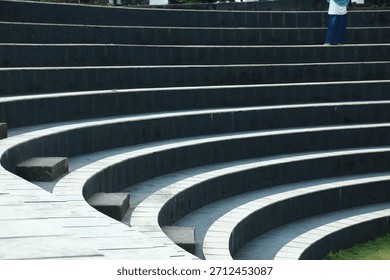 An abstract pattern of tiered circular steps or tribunes, consisting of light square stone tiles separated by dark gaps, with a green grass background to the side.