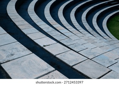 An abstract pattern of tiered circular steps or tribunes, consisting of light square stone tiles separated by dark gaps, with a green grass background to the side.