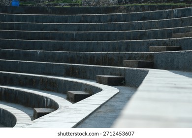 An abstract pattern of tiered circular steps or tribunes, consisting of light square stone tiles separated by dark gaps, with a green grass background to the side.