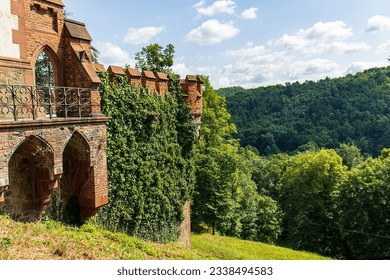Abstract photo of the side of a Gothic castle. Corner of the Gothic castle building. Terrace with decorative railings, Gothic arches, red bricks. Forest, nature. grass. Blue sky with white clouds.
