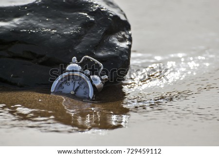 an abstract shot of a clock leaning on a rock on the beach, sinking in sand and ocean water. 