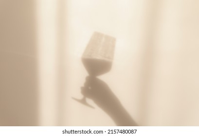 Abstract silhouette shadows hand holding glass of wine in the rays the sun reflected on wall. Blurry and out of focus.