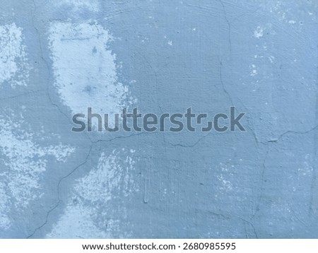 An abstract texture of a weathered wall. The surface is a faded blue with prominent cracks and large, mottled patches of lighter, almost white, paint. This image provides a distressed, rustic.