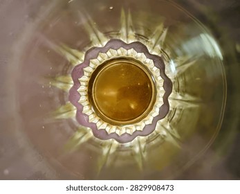 Abstract top-down view through the bottom of a crystal glass. The glass has intricate starburst pattern with scalloped edges and creates a golden amber reflection in the center. Soft blurry background with warm tones. The image shows refraction, light patterns, and elegant glass texture. Perfect for concepts of abstract art, luxury, beverage, tea, whiskey, wine, background texture, pattern, and elegant design. High detail macro photography.