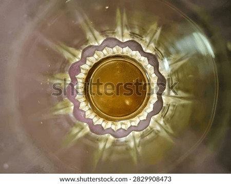 Abstract top-down view through the bottom of a crystal glass. The glass has intricate starburst pattern with scalloped edges and creates a golden amber reflection in the center. Soft blurry background with warm tones. The image shows refraction, light patterns, and elegant glass texture. Perfect for concepts of abstract art, luxury, beverage, tea, whiskey, wine, background texture, pattern, and elegant design. High detail macro photography.