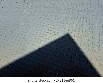 Abstract triangular shadow cast on textured metal floor under sunlight. Minimal composition with strong contrast, geometry, and industrial background.