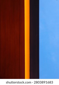 Abstract vertical straight line intersection of brown wood panel material warm yellow linear LED light and building wall against blue sky.