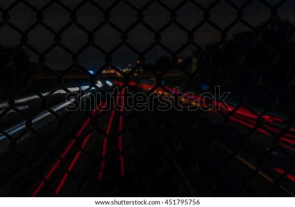 Abstract view of light streaks on busy highway.\
Light painting at night. Cars racing. Fast cars and light streaks.\
Chain link fence view. Abstract art and design. Industrial design\
and art. Highway.