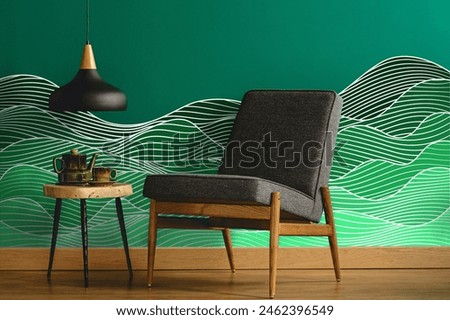 Abstract wave pattern wall decal or wall sticker on a teal green wall with a gray upholstered chair by a ceiling lamp over a wooden side table in a minimalist living room interior.