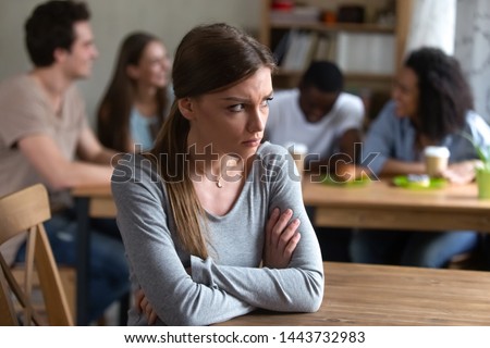 Abused upset offended by friends young caucasian woman sitting alone separately from group of happy laughing people, sad introvert feeling lonely, suffering from bullying, low self-esteem concept.