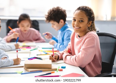 Academic Concept. Smiling junior african american school girl sitting at desk in classroom, writing in notebook, posing and looking at camera. Group of classmates studying in the background - Shutterstock ID 1903221799