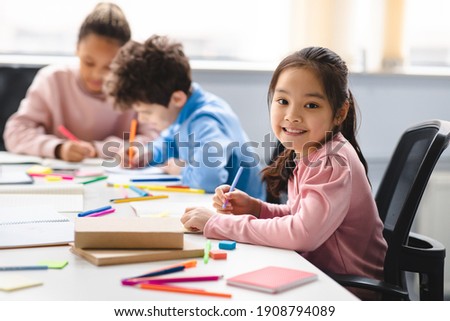 Academic Concept. Smiling junior asian school girl sitting at desk in classroom, writing in notebook, posing and looking at camera. Group of diverse classmates studying in the background