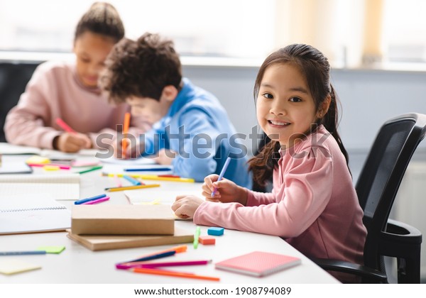 Academic\
Concept. Smiling junior asian school girl sitting at desk in\
classroom, writing in notebook, posing and looking at camera. Group\
of diverse classmates studying in the\
background