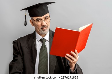 Academician, Professor, Prof Is Wearing A Cap With Tassel And Black Gown. Senior Educator Holding Red Folder. Doctor Of Philosophy, Learned Man From A University Or College.