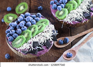 Acai Smoothie Bowl With Fresh Kiwi, Frozen Blueberries, Organic Coconut And Dark Chocolate Pieces With Wooden Spoons Served In Coconut Bowls Over A Rustic Table. Shot From Above / Overhead.