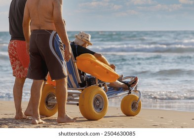 Accessible beach wheelchair helps individual enjoy the ocean at sunset. A beach wheelchair with large yellow wheels supports a person sitting in it, as two individuals stand nearby, enjoying the sea - Powered by Shutterstock