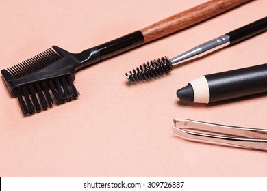 Accessories for care of brows: eyebrow pencil, tweezers, brush and comb on peach colored textured surface. Eyebrow grooming tools