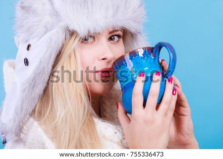 Accessories and clothes for cold days, fashion concept. Blonde woman in winter warm furry hat drinking hot drink from mug. Blue background.