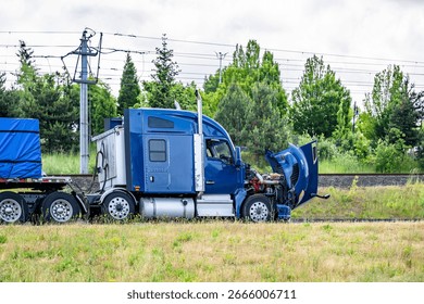 Accidentally broken down while driving  blue big rig semi truck with open hood and loaded flat bed semi trailer stands on the highway shoulder waiting for mobile repair service road assistant