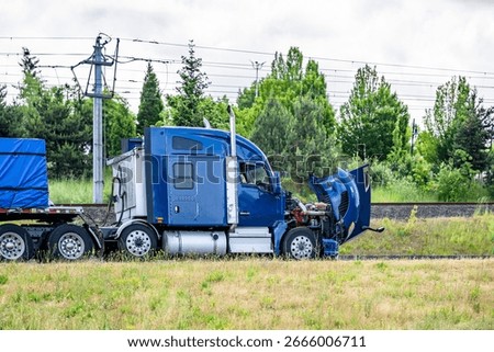 Accidentally broken down while driving  blue big rig semi truck with open hood and loaded flat bed semi trailer stands on the highway shoulder waiting for mobile repair service road assistant