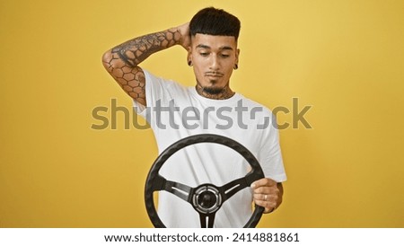 Accident-prone young latin man with tattoo struggling, steering wheel drive mishap on isolated yellow background portraying serious expression
