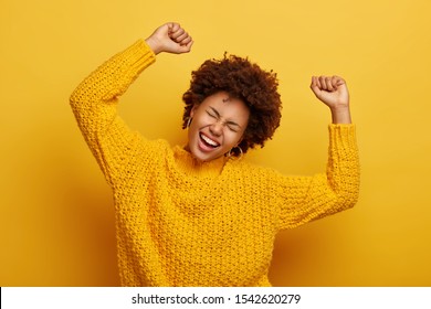 Accomplishment and success concept. Joyful Afro woman raises arms, feels glad and relaxed, tilts head, dressed in casual knitted jumper, laughs from happiness, celebrates victory, isolated on yellow
