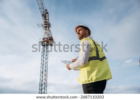 According to a plan. Construction site worker is against building crane and sky background.