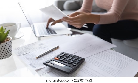 Accountant woman pay bills online use e-bank app, calculating household finances or taxes sit on sofa at home office. Family expenditures management, close up focus on calculator and heap of receipts