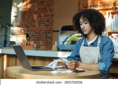 Accounting, Taxes And Bill Payment During COVID-19 Lockdown. Serious African American Female Bartender Manager In Apron Works With Financial Accounts And Bookkeeping In Cafe, Empty Space