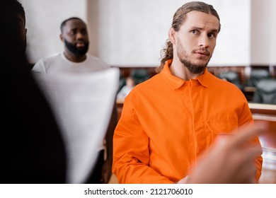 Accused Man In Jail Uniform Looking At Blurred Prosecutor Pointing With Finger In Courtroom