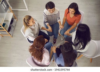 Achieving Goals And Overcoming Problems Together. Happy Women Sitting In Circle And Holding Hands In Support Group Meeting With Coach Or Therapy Session With Psychotherapist. From Above, High Angle