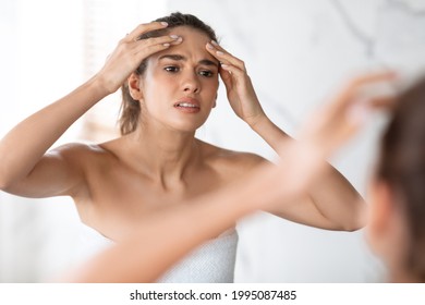 Acne Issue. Frustrated Lady Squeezing Pimple On Forehead Looking At Reflection In Mirror Standing In Modern Bathroom Indoors. Facial Skin Problems, Pimples Abscess Issue Concept. Selective Focus