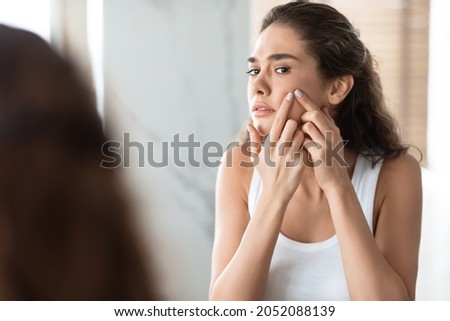 Acne Issue. Unhappy Female Squeezing Pimples On Cheek Looking At Mirror In Bathroom Indoor. Facial Skin Problems, Treatment And Care For Blemish-Prone Skin Concept. Selective Focus