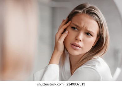 Acne prone skin concept. Sad lady looking at mirror and checking her face skin, standing in bathroom interior, selective focus on her reflection - Shutterstock ID 2140809513