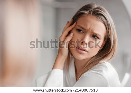 Acne prone skin concept. Sad lady looking at mirror and checking her face skin, standing in bathroom interior, selective focus on her reflection