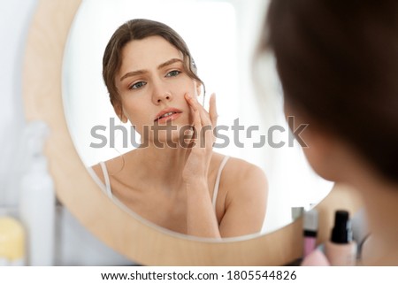 Acne prone skin concept. Upset young woman looking at mirror at home, checking her face skin