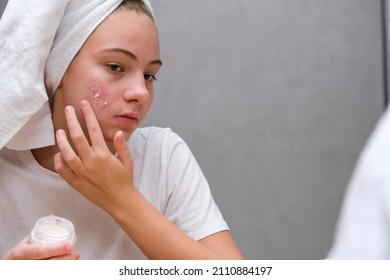 Acne. A teenage girl applying acne medication on her face in front of a mirror. Care for problem skin - Shutterstock ID 2110884197
