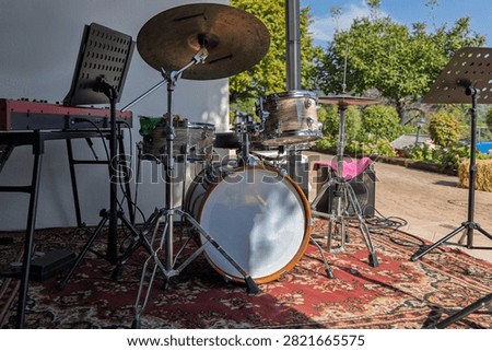 An acoustic drum kit is centered between a red stage keyboard and two music stands on a patterned rug. Cables, cymbals and the bright garden beyond suggest a relaxed small-band performance in a home patio setting. The empty instruments emphasize preparation before musicians begin to play.