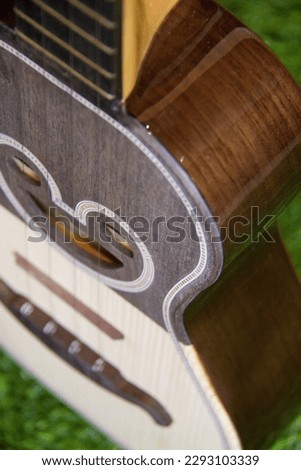 Acoustic ukulele made of wood in two colors, with a heart soundhole, resting on a wooden bench and on artificial grass, with a background of acoustic guitars hanging on a wooden wall