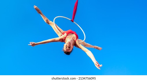 acrobat athletic, young graceful gymnast performing aerial exercise in the air ring outdoors on sky background. flexible woman in red suit performs circus artist dancing on hoop upside down. low angle