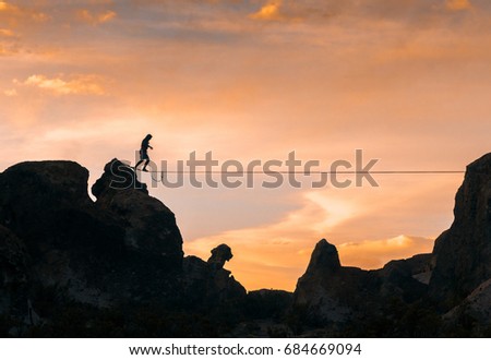 An acrobat walking the slack line between two mountains at dusk with an orange sky in the background.