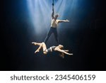 An acrobatic performance featuring an aerial straps duet: a man gracefully hangs by one hand, while a woman lies extended at his feet. They are bathed in an ethereal blue and white glow