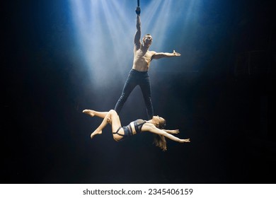An acrobatic performance featuring an aerial straps duet: a man gracefully hangs by one hand, while a woman lies extended at his feet. They are bathed in an ethereal blue and white glow