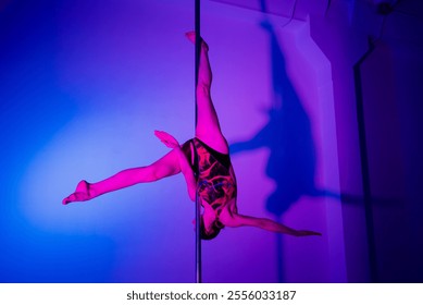 Acrobatic pole dance. Grace and strength, professional dancer. A woman is practicing gymnastics on a pole in the neon light of the studio. - Powered by Shutterstock